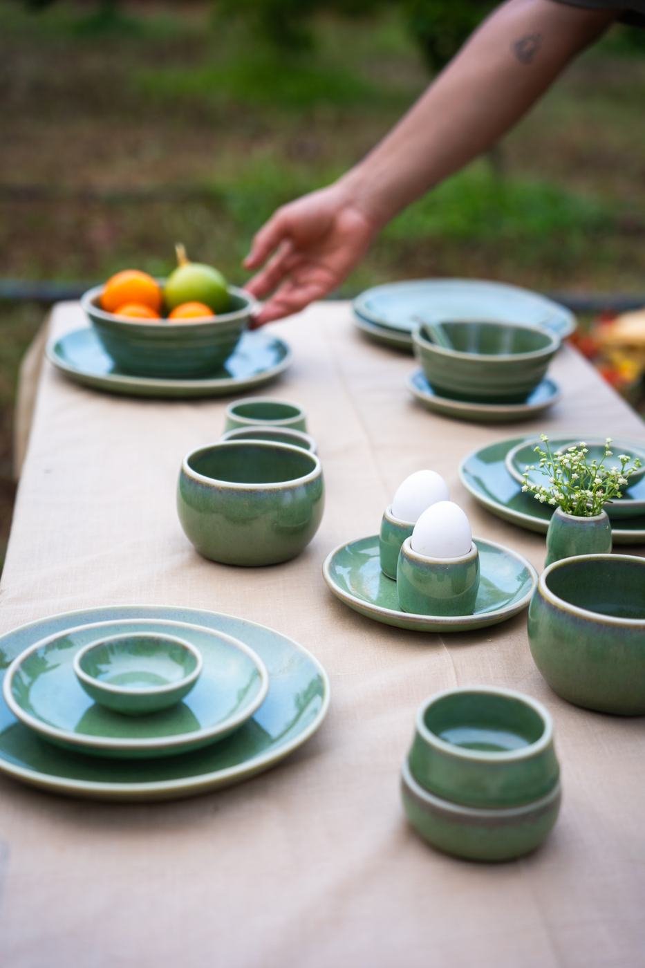 A hand reaching across a table set with sage green Nugu bowls, plates and egg cups