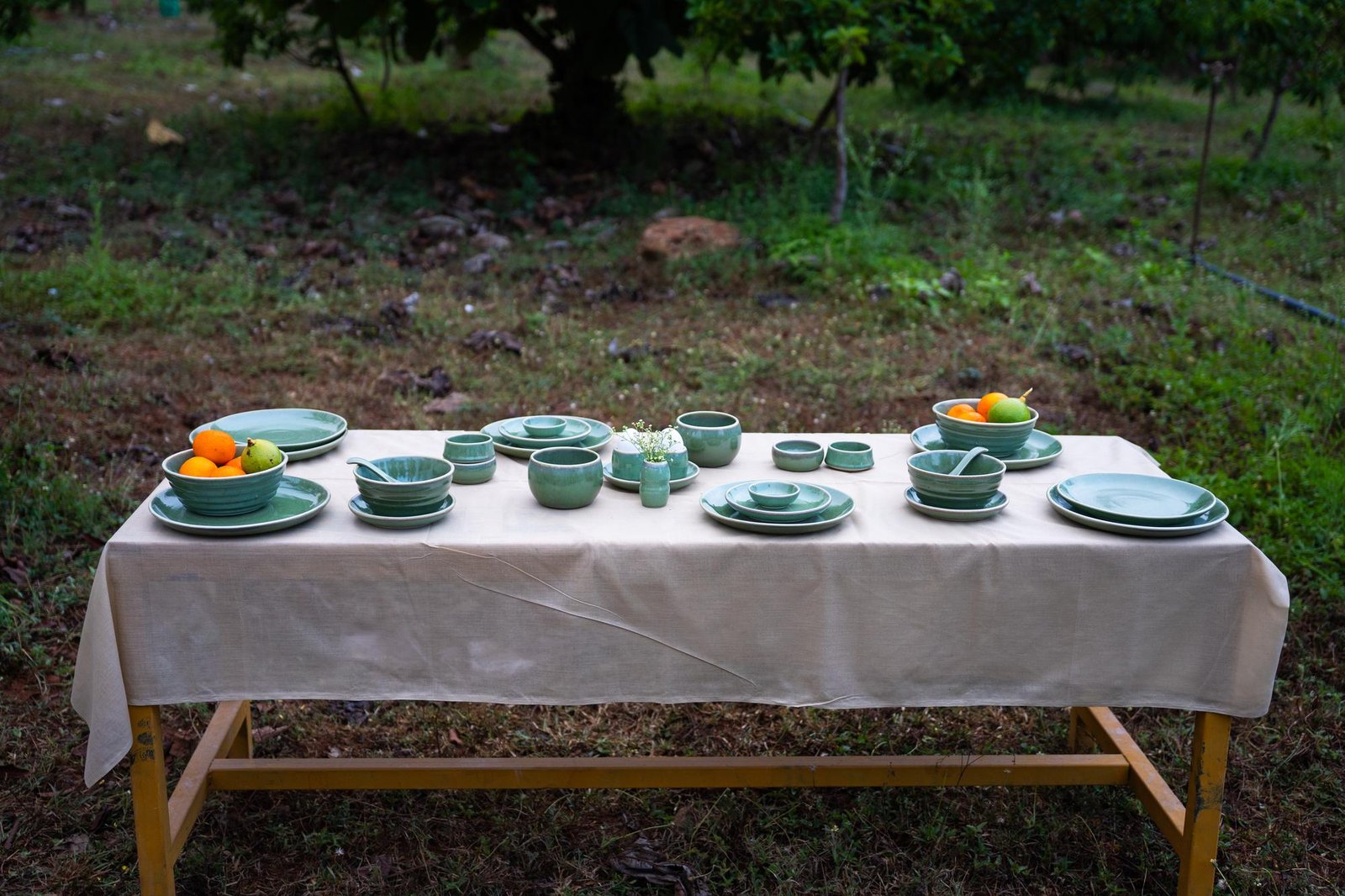 A long outdoor table set with a full sage green Nugu dinner service, fruit bowls and egg cups